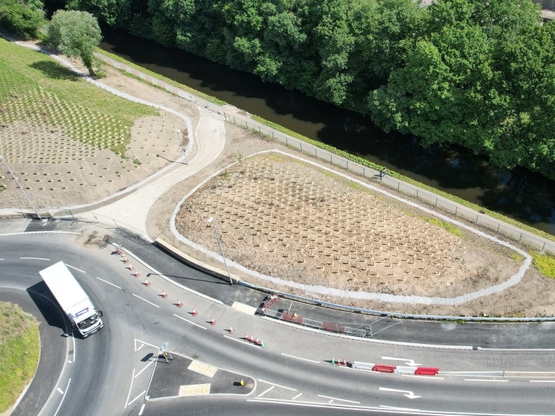 A lorry navigates one segment of the roundabout near the turnoff for the bridge. Recently planted young trees are spaced out evenly between the road and the canal. 